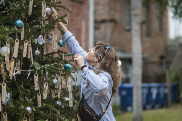 A woman reaches up to hang a wooden tag ornament on a decorated outdoor Christmas tree, which is covered with handwritten tags and blue and silver baubles.