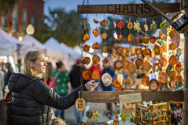A woman in a black jacket browses colorful, handcrafted ornaments hanging from chains at an outdoor market booth, with white tents and people in the background.