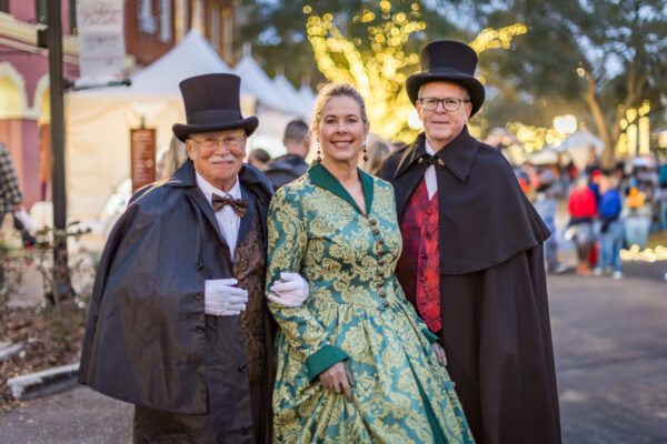 Three adults in Victorian-era costumes, including top hats and a green patterned dress, stand arm-in-arm at an outdoor event with festive lights and blurred people in the background.