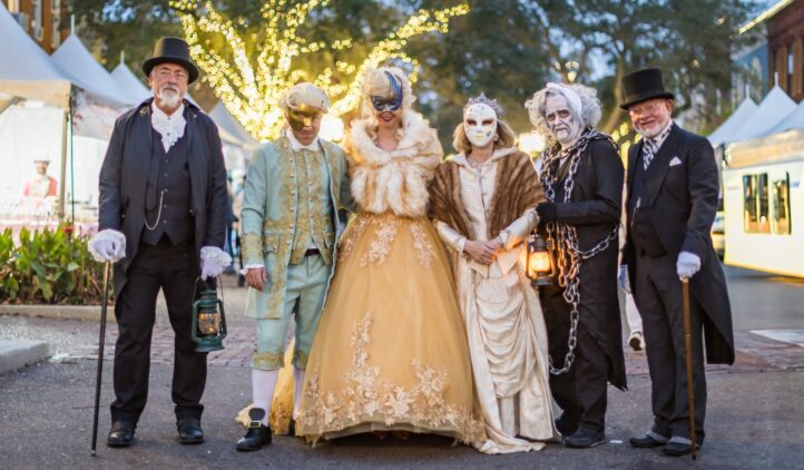 Six people in elaborate historical and masquerade costumes stand together outdoors at dusk, with festive lights and tents in the background. Some hold lanterns and wear masks, creating a festive and mysterious atmosphere.