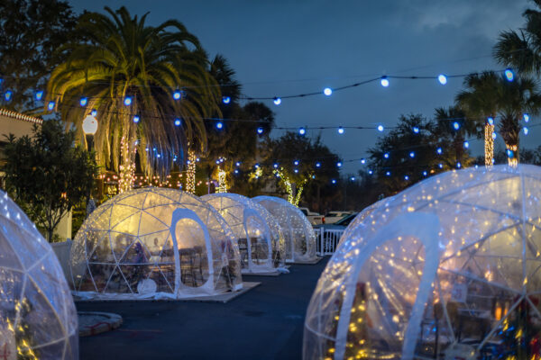 A nighttime outdoor dining area features transparent igloo tents decorated with string lights, surrounded by palm trees wrapped in lights, and blue bulbs hanging overhead, creating a festive atmosphere.