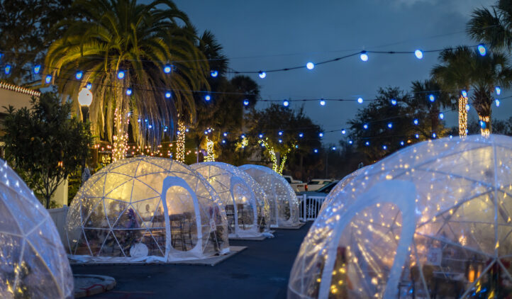 A nighttime outdoor dining area features transparent igloo tents decorated with string lights, surrounded by palm trees wrapped in lights, and blue bulbs hanging overhead, creating a festive atmosphere.