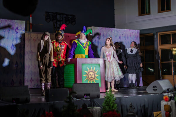 A group of five people in colorful costumes perform on a stage decorated with a sun emblem and festive backdrop. They appear to be acting out a scene, with dramatic expressions and theatrical lighting.