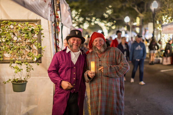 Two men in festive costumes stand smiling at a holiday market; one wears a purple coat and top hat, the other a plaid nightshirt and red cap, holding a candle. The background is decorated with lights and people.