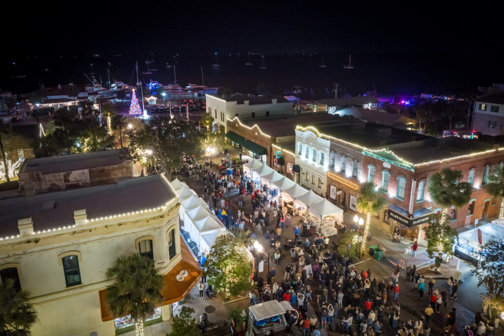 Aerial view of a festive nighttime street market with crowds, white vendor tents, and historic buildings decorated with string lights near a waterfront with boats and a lit Christmas tree.