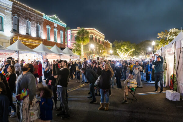 A large crowd gathers at a lively outdoor street festival at night, surrounded by white tents, food vendors, and illuminated historic buildings with festive lights. Some people are standing, while others are seated or taking photos.