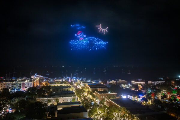 A night scene of a city by the water, with colorful drone lights in the sky forming a festive image of Santa on a sleigh under stars. The streets below are illuminated with holiday lights.