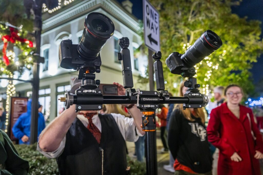 A person holds a large dual camera rig with two cameras mounted side by side on a busy street decorated with holiday lights, while people walk and gather in the background.