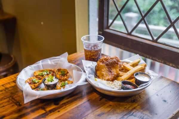 Two plates of food on a wooden table by a window: one with loaded potato skins and dips, the other with fish, fries, coleslaw, and sauces. A plastic cup of soda sits behind the plates.