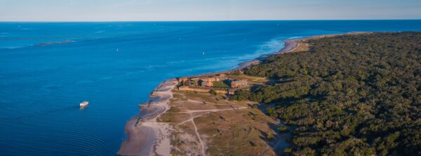 Aerial view of a historic fort surrounded by dense forest, located on the edge of a sandy shoreline with blue ocean water. A small boat is visible near the coast under a clear sky.