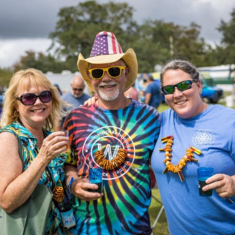 Three people smile at an outdoor event. The man in the center wears a tie-dye shirt, American flag hat, and yellow sunglasses. The two women beside him hold drinks and wear leis made of pretzels. There are people and trees in the background.