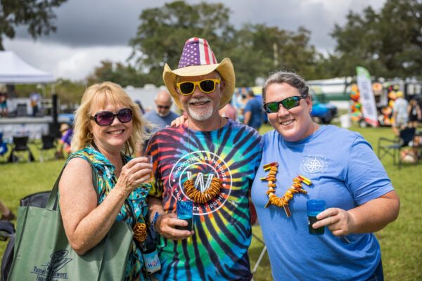 Three people smile at an outdoor event. The man in the center wears a tie-dye shirt, American flag hat, and yellow sunglasses. The two women beside him hold drinks and wear leis made of pretzels. There are people and trees in the background.