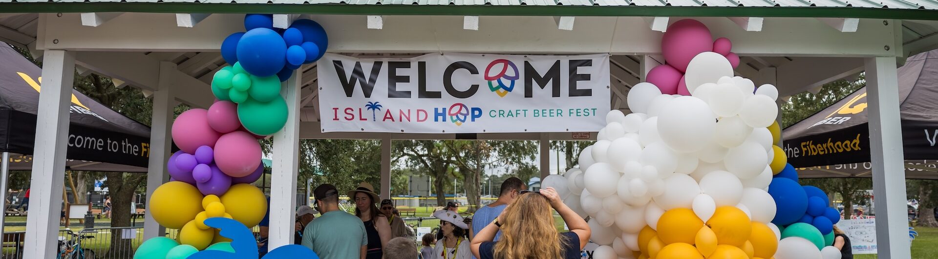 People gather under a gazebo decorated with colorful balloons and a sign reading Welcome Island Hop Craft Beer Fest. Palm tree and peace symbols are part of the decor, and trees and tents are visible in the background.