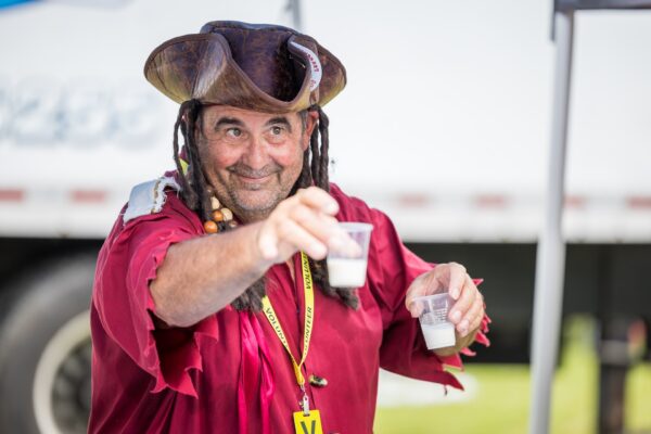A smiling man in a pirate hat and red shirt holds out two small cups with a light-colored drink, standing outdoors with a blurred background.