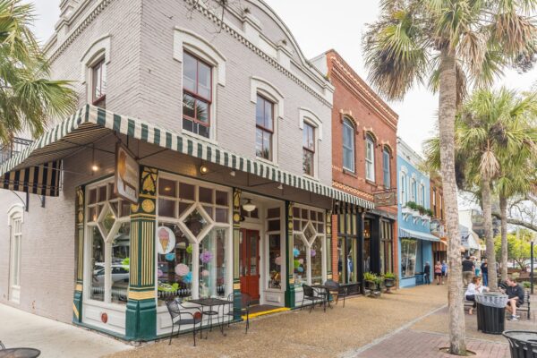 Street view of colorful shops and cafes with large windows and striped awnings on a tree-lined sidewalk with outdoor seating and people relaxing under palm trees.
