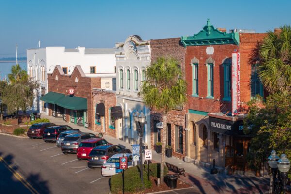 A row of historic brick and stucco buildings lines a sunny street with parallel-parked cars in front. Palm trees and vintage storefronts create a charming small-town atmosphere under a clear blue sky.