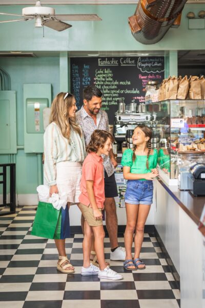 A smiling family of four stands together inside a cafe, with two children looking at pastries in a display case. The parents stand behind them on a black-and-white checkered floor. The cafe menu is visible in the background.