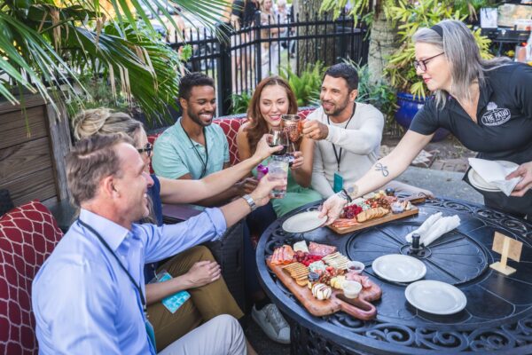 A group of five people sit around an outdoor table, smiling and raising drinks for a toast. A server stands nearby, serving food. The table is set with charcuterie boards, fruit, and snacks. Lush plants surround the patio area.