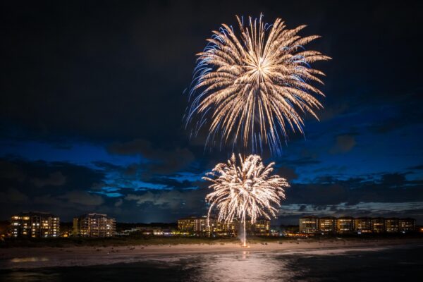 Fireworks light up the night sky above a beach, with several bursts visible. In the background, illuminated buildings line the shore under a deep blue sky with scattered clouds.