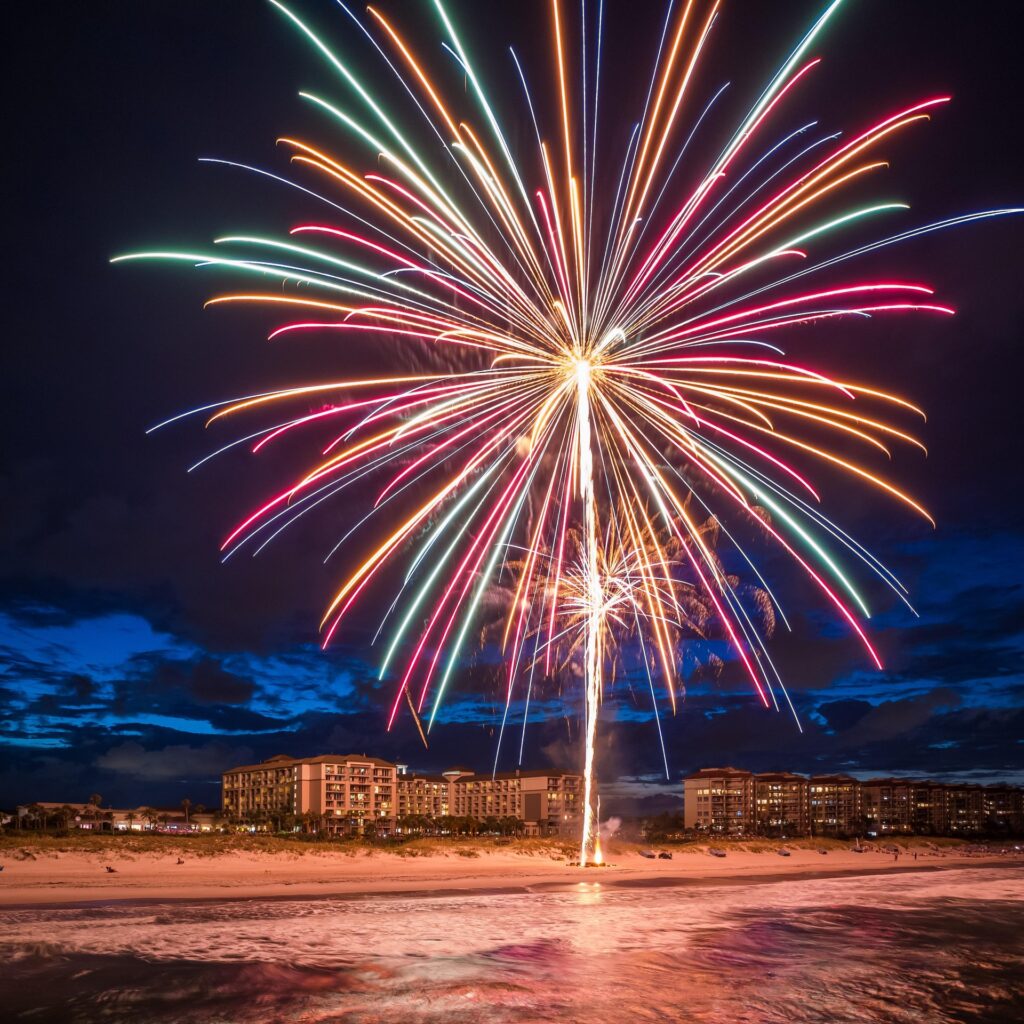 Colorful fireworks burst over a beach at night, illuminating the sky and reflecting on the water, with several buildings and a dark blue, cloudy sky in the background.