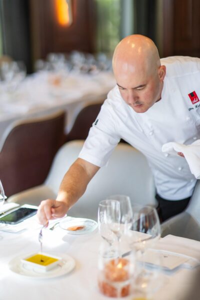A chef in a white uniform delicately garnishes a dish at a set dining table, surrounded by wine glasses and elegant place settings in a restaurant.