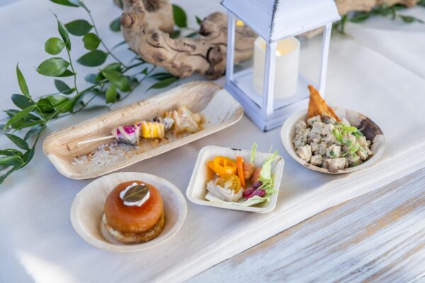 A selection of gourmet appetizers in small wooden dishes, arranged on a white tablecloth with green leaves, a white lantern with a candle, and a decorative wooden branch in the background.