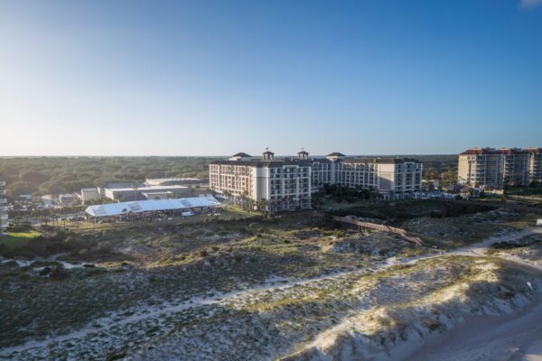 Aerial view of a large beachfront resort surrounded by greenery and sand dunes under a clear blue sky, with several buildings and tents visible on the property.