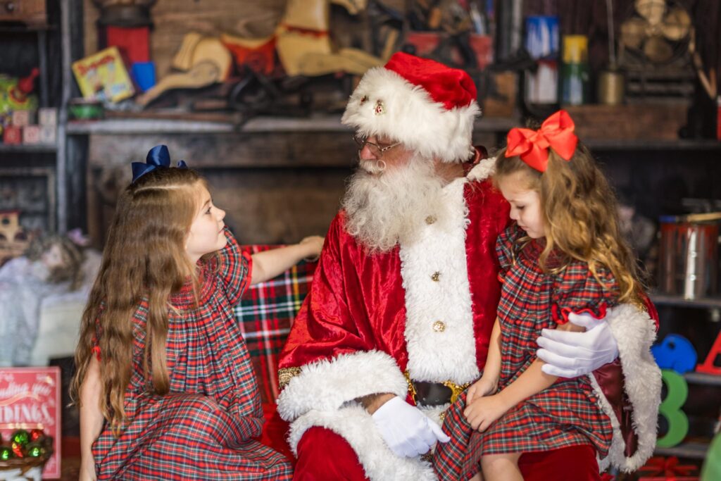 Santa Claus sits between two young girls wearing matching plaid dresses and festive bows, as they talk and smile in a cozy, decorated room with holiday toys and decorations in the background.