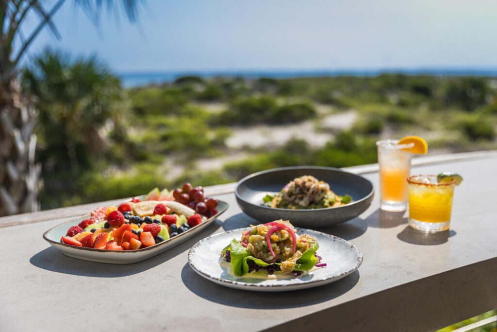 Plates of fresh fruit, salad, and seafood sit on an outdoor table, with two citrus drinks beside them. The background shows greenery, palm trees, and a view of the ocean under a sunny sky.