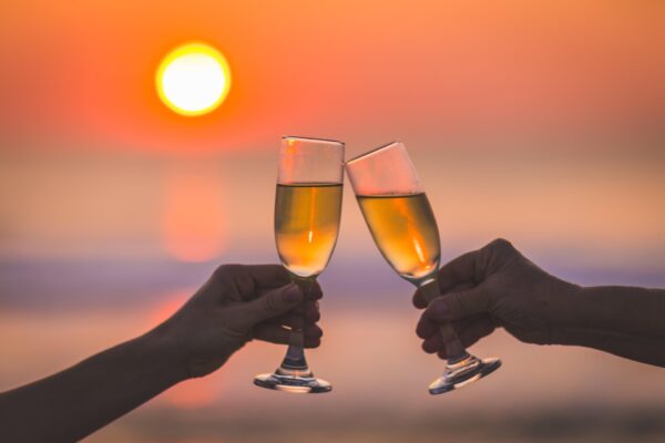 Two hands clink champagne glasses together with a vibrant orange sunset over the ocean in the background, celebrating or toasting at the beach.
