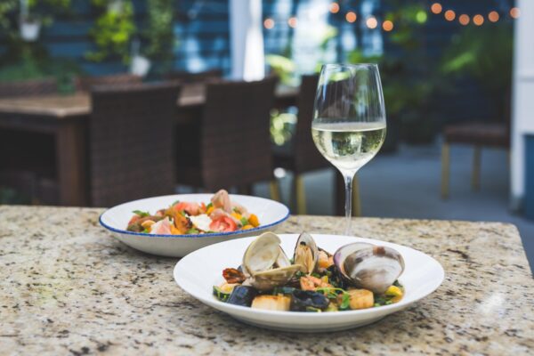 A glass of white wine sits on a granite table next to two plates of seafood dishes, with an outdoor dining area and string lights blurred in the background.