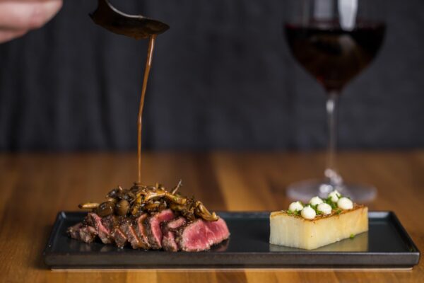 A hand pours sauce over sliced rare steak topped with mushrooms, served next to a rectangular piece of potato garnished with small white dots, with a glass of red wine in the background.