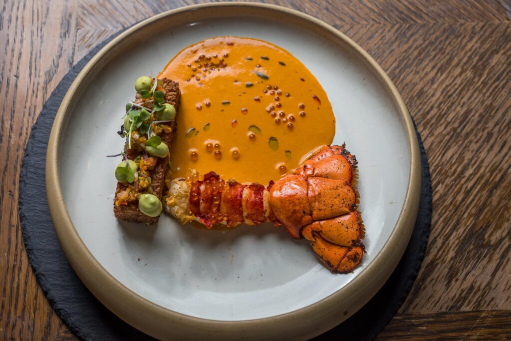 A plated gourmet dish featuring a lobster tail, a slice of toasted bread topped with green sauce and microgreens, and a pool of creamy orange sauce garnished with droplets, on a round white plate.