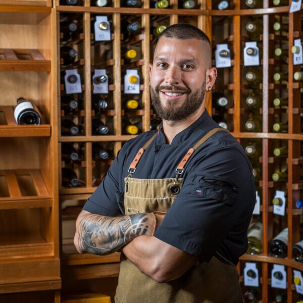 A smiling man with a beard and tattoos, wearing a black shirt and brown apron, stands with arms crossed in front of shelves filled with bottles of wine.