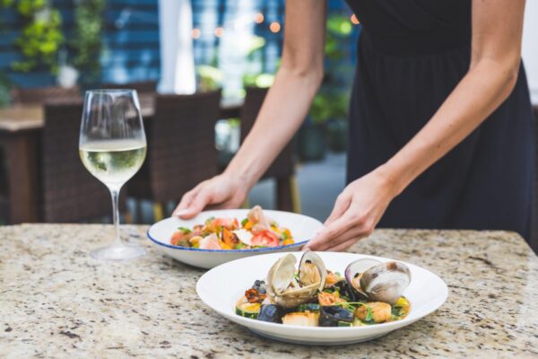 A person in a black dress sets down a plate of seafood pasta on a marble table next to a glass of white wine and another dish, in an outdoor dining area.