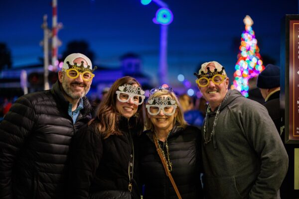 Four people wearing festive Hello 2024 glasses and crowns smile together outdoors at night. They are dressed warmly, and colorful holiday lights and a Christmas tree appear in the blurred background.