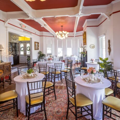 Elegant dining room with round tables covered in white cloths, gold cushioned chairs, floral centerpieces, and large windows letting in natural light. Ornate decor and a red ceiling complete the inviting atmosphere.