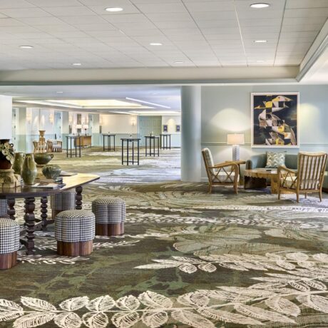 Spacious hotel lobby with patterned carpet, round table with decor, stools, lounge chairs, and modern art on light blue walls. Large windows bring in natural light.