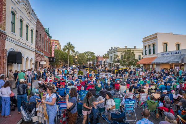 A large crowd gathers with chairs in a historic downtown street surrounded by brick buildings and shops, enjoying an outdoor event under a clear blue sky.