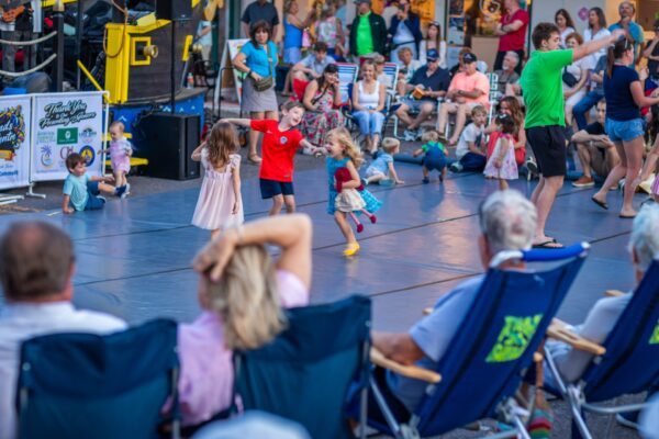 Children dance and play on a mat in front of a seated crowd at an outdoor event, while adults watch and relax in folding chairs. The atmosphere is lively and cheerful.