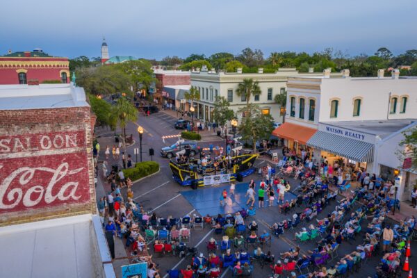 A lively outdoor event features a boxing ring surrounded by a crowd seated in folding chairs on a downtown street, with historic buildings, shops, and trees visible under a clear evening sky.