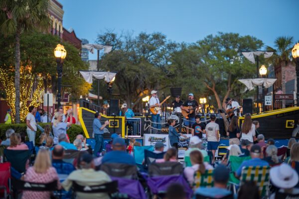 A band performs on an outdoor stage styled like a ship in a town square, as a crowd seated on lawn chairs watches during the evening. Strings of lights are wrapped around a nearby tree, and buildings and greenery are visible in the background.