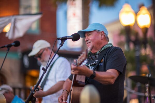 Two older men play guitars and sing into microphones at an outdoor event. One wears a blue cap and black shirt, smiling as he performs, with a blurred background of buildings and streetlights at dusk.