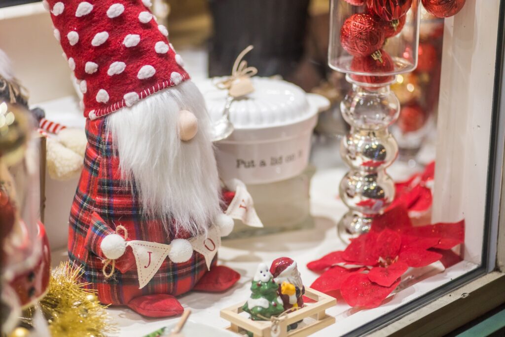 A festive holiday display features a gnome doll in a red plaid outfit and spotted hat, holding a JOY banner. Theres a small sled with a mouse figurine, red ornaments, poinsettia flowers, and a white jar in the background.