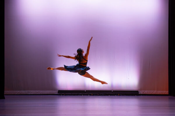 A dancer in a dark dress performs a high split leap on stage, with arms extended and dramatic lighting casting soft purple and pink tones on the background.