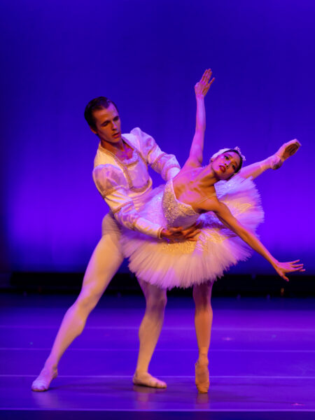 Two ballet dancers perform on stage in elegant costumes. The male dancer supports the female dancer, who wears a white tutu and tiara, as she poses gracefully with arms extended. The background is lit with purple light.