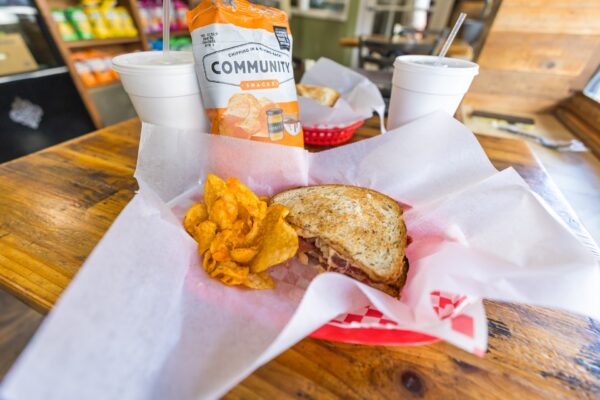 A deli sandwich and potato chips are served in a red basket lined with white paper, next to a bag of COMMUNITY Snacks chips and two white drink cups on a wooden table in a casual eatery.