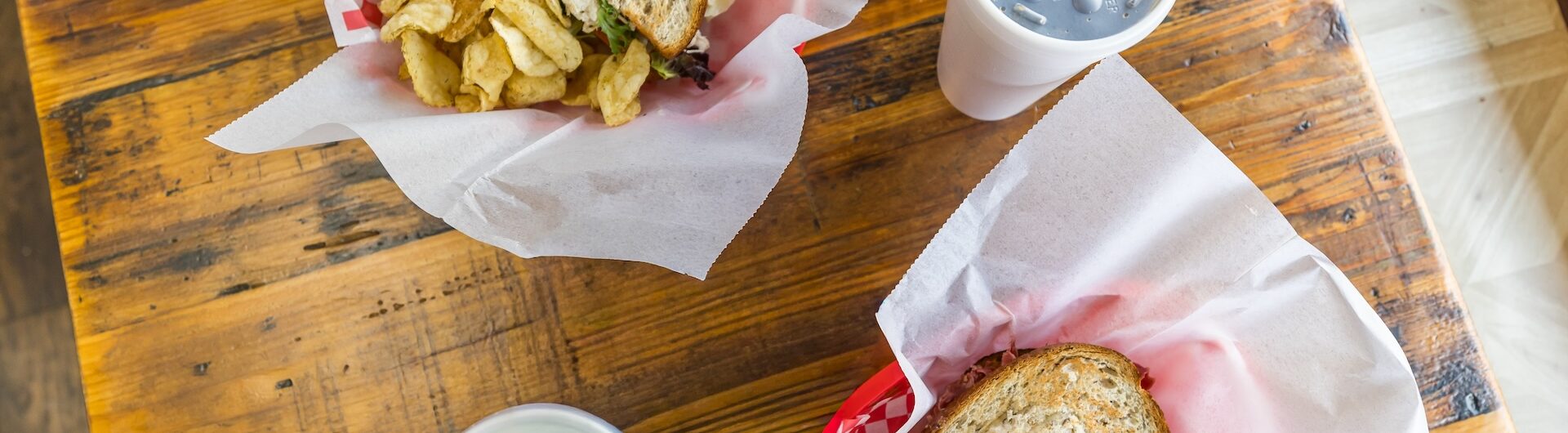 Two baskets with sandwiches and chips sit on a wooden table, each accompanied by a paper cup with a lid and straw. One basket has potato chips; the other has orange chips, possibly barbecue-flavored.
