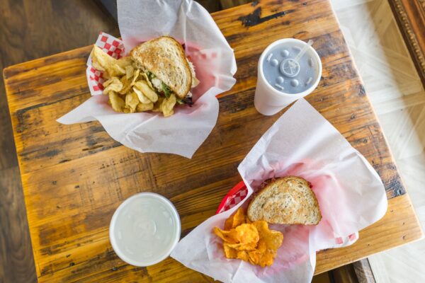 Two baskets with sandwiches and chips sit on a wooden table, each accompanied by a paper cup with a lid and straw. One basket has potato chips; the other has orange chips, possibly barbecue-flavored.
