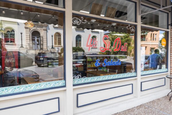 Street view of a deli storefront with large windows displaying the name 4th St. Deli & Salads in red and blue lettering. Reflections of nearby buildings are visible on the glass.